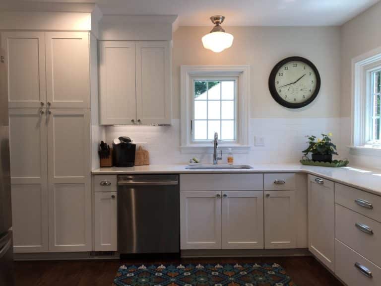 White kitchen with cabinets, sink under two windows, wall clock, plants, and a colorful rug on the floor.
