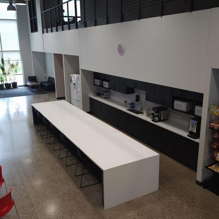 Modern office kitchen area with a long white table, black stools, appliances, and snacks on shelves.