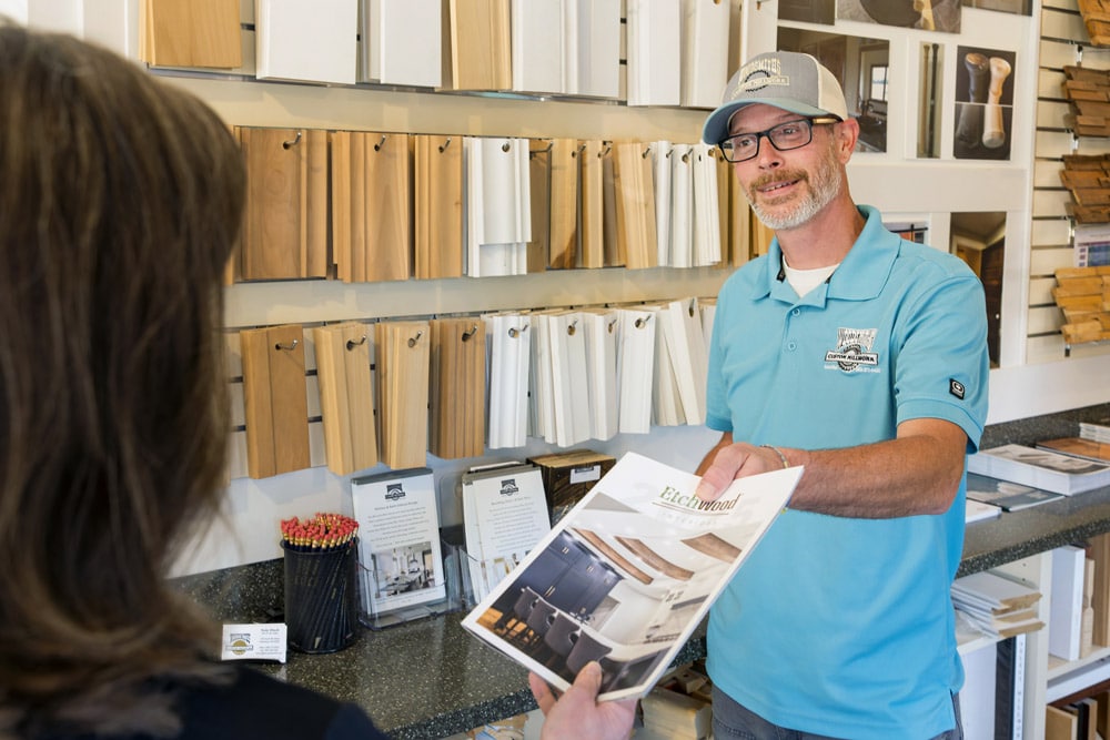 A man in a blue polo shows a brochure to a woman in a showroom with wood samples on the wall.