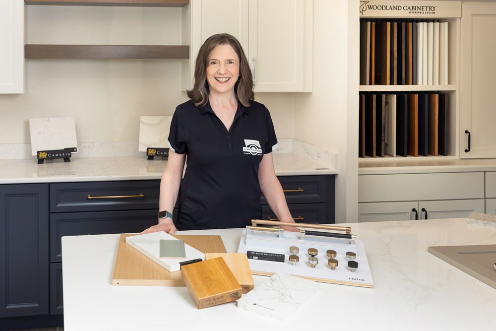 Smiling woman stands at a counter with cabinet samples, hardware, and design materials in a showroom.