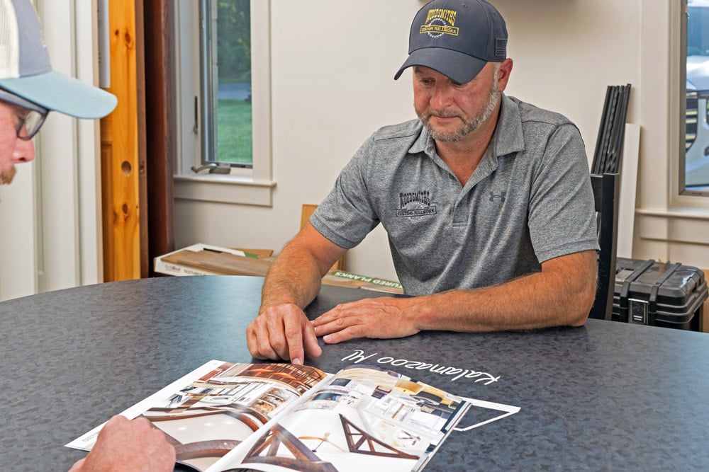 Two men review a home design catalog at a round table in a well-lit office.
